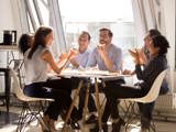 Sex styrelseledamöter skrattar och delar på pizzor runt ett bord i en lägenhet.
